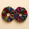 Two Afrocentric fabric scrunchies with vibrant geometric and floral patterns on a beige background.
